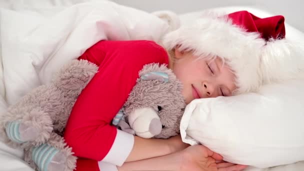 Dziecko śpi przed świętami. Mała dziewczynka w kapeluszu Mikołaja i świąteczna piżama leżąca w białym łóżku przytulająca misia w domu. Nowy Rok i święta. poranne szczęście. — Wideo stockowe