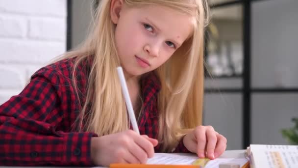 4k écolière blonde étudiant à la maison faisant des devoirs scolaires. Regarder et sourire à la caméra. Livres de formation et cahiers sur table. Enseignement à distance en ligne. — Video