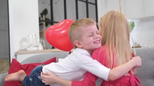 Kleine jongen knuffelen geliefde meisje vieren Valentijnsdag met rode hartvorm ballonnen thuis. Langzame beweging. — Stockvideo