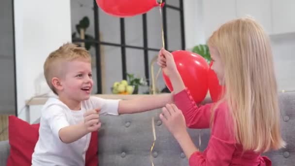 Μικρό αγόρι αγκαλιάζει αγαπημένο κορίτσι Γιορτάζοντας την Ημέρα του Αγίου Βαλεντίνου με κόκκινα μπαλόνια σχήμα καρδιάς στο σπίτι. Αργή κίνηση. — Αρχείο Βίντεο