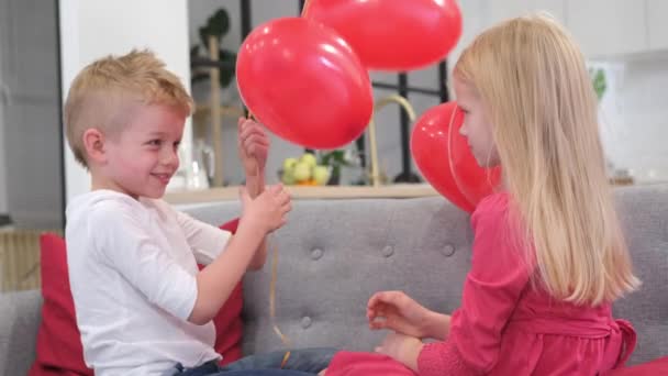 Little boy gives his beloved girl Valentines Day red heart shapes balloons. Happy smiling blonde kids celebrating Valentines day. Slow motion Stock Video