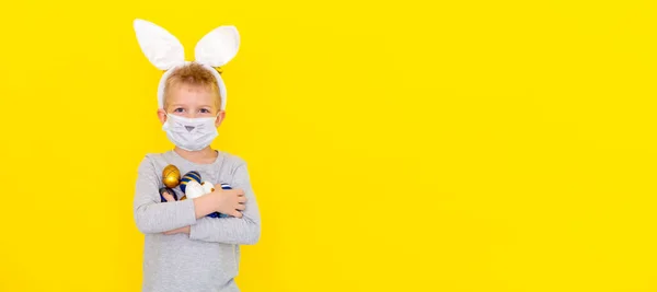 Banner copyspace Ragazzo in coniglio orecchie da coniglio sulla testa e maschera protettiva con uova colorate su sfondo giallo studio. Allegro pazzo sorridente bambino felice. Covid bambino pasquale — Foto Stock