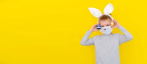 Junge mit Hasenohren auf dem Kopf und Schutzmaske mit bunten Eiern auf gelbem Studiohintergrund. Fröhlich verrückt lächelndes glückliches Kind. Covides Osterkind — Stockfoto