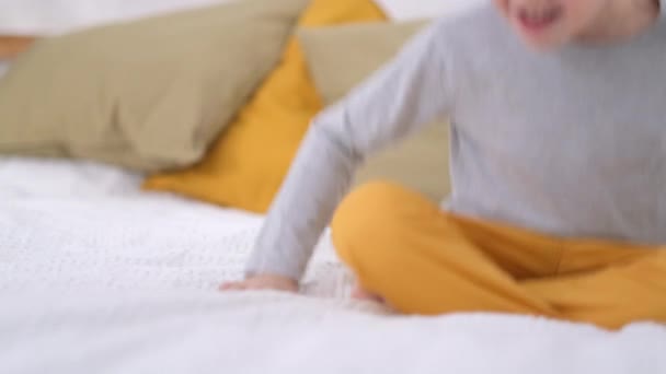 Blonder Junge springt und fällt auf weißes Bett. Aufgeregte kaukasische Kind in gelben Hosen Spaß zu Hause genießen Lachen beim lustigen aktiven Spiel im Schlafzimmer — Stockvideo