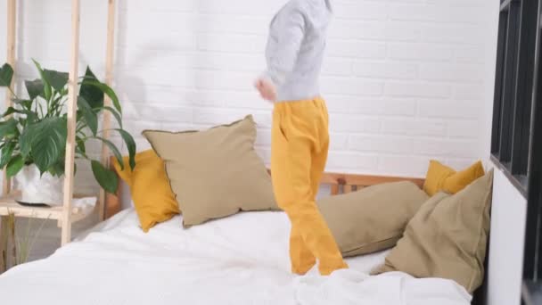 Ξανθό αγόρι που πηδάει στο λευκό κρεβάτι. Ενθουσιασμένος καυκάσιος παιδί με κίτρινο παντελόνι που διασκεδάζει στο σπίτι απολαύσετε το γέλιο παίζοντας αστείο ενεργό παιχνίδι στην κρεβατοκάμαρα — Αρχείο Βίντεο