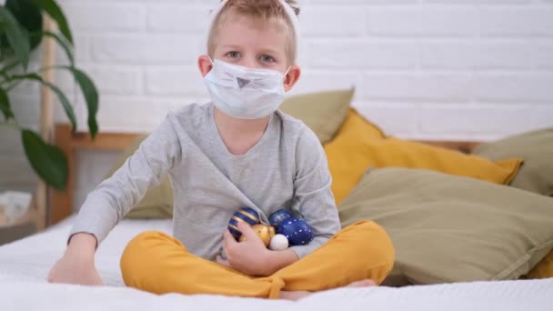 Paasjacht. Jongen in konijn konijnenoren op het hoofd en beschermende masker het verzamelen van kleurrijke eieren plezier hebben op bed thuis. — Stockvideo