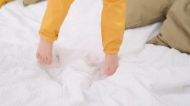 Beyaz yatakta zıplayarak bacaklarını kapat. Heyecanlı sarı pantolonlu beyaz çocuk evde eğleniyor. Yatak odasında eğlenceli oyunlar oynuyor.