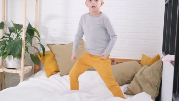 Blondyn skaczący na białym łóżku. Podekscytowane dziecko kaukaskie w żółtych spodniach zabawy w domu cieszyć się śmiechem gry śmieszne aktywne gry w sypialni — Wideo stockowe