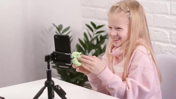 フォロワーと話す小さな女の子ブロガーのインフルエンサーは、緑のスライムでライブストリーミング再生します。横に伸びる傾向のある液体おもちゃ。自宅でスマートフォンの画面を探している子供。スローモーション — ストック動画