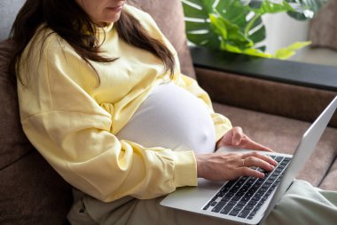 Evden internet üzerinden çalışan çekici hamile bir genç bayan. Kadın ellerini dizüstü bilgisayarın üzerine koy ve karnında büyük bir hamilelik var.