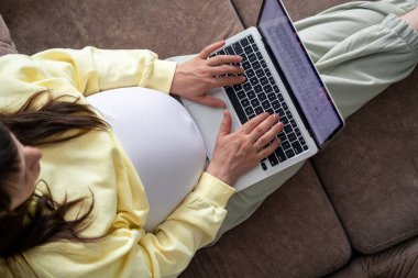 Evden internet üzerinden çalışan çekici hamile bir genç bayan. Kadın ellerini dizüstü bilgisayarın üzerine koy ve karnında büyük bir hamilelik var.
