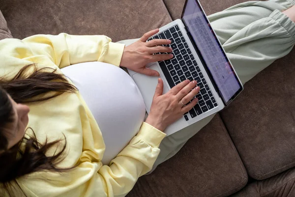 Evden internet üzerinden çalışan çekici hamile bir genç bayan. Kadın ellerini dizüstü bilgisayarın üzerine koy ve karnında büyük bir hamilelik var.