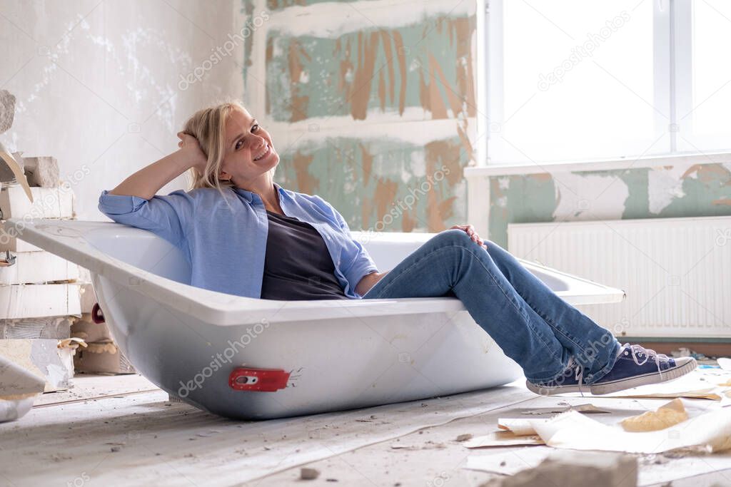 Renovation apartment. Creative story young happy woman sits in bathtub in the middle of the room. Empty walls, repairs house with their own hands