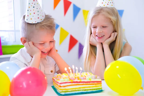 금발의 두 남자 아이들 과 여자 아이들 이 생일 파티에서 촛불을 켜고 무지개 케이크에 촛불을 불며 즐겁게 놀고 있습니다.. — 스톡 사진