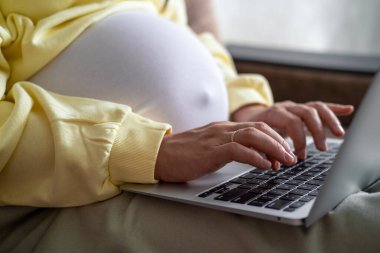 Evden internet üzerinden çalışan çekici hamile bir genç bayan. Kadın ellerini dizüstü bilgisayarın üzerine koy ve karnında büyük bir hamilelik var.
