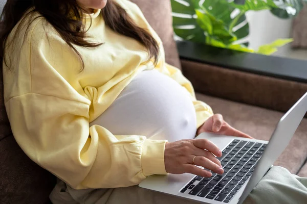Evden internet üzerinden çalışan çekici hamile bir genç bayan. Kadın ellerini dizüstü bilgisayarın üzerine koy ve karnında büyük bir hamilelik var.