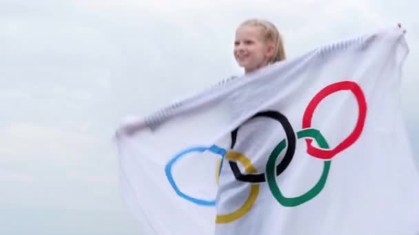 4k. Menina loira acenando bandeira dos Jogos Olímpicos ao ar livre sobre céu nublado pôr do sol. Crianças fã de esportes. Jogos Olímpicos de Verão. 23.03.2020 St.Petersburg Rússia. — Vídeo de Stock