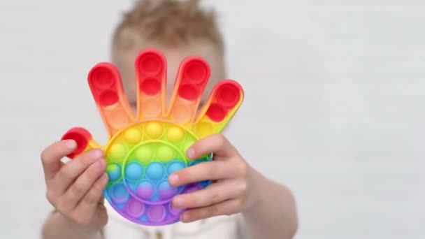 Retrato chico rubio con pop forma de mano de juguete sensorial. Chico presiona colorido arco iris burbujas de silicona suave y blanda sobre el fondo de ladrillo blanco. Alivio de estrés y ansiedad. De moda inquieto juego — Vídeos de Stock