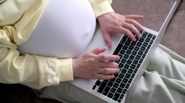 4k Top view Bekleyen Anne evden internet üzerinden çalışıyor. Hamile genç kadın ellerini dizüstü bilgisayarına koyup karnına büyük bir hamilelik geçirmiş.