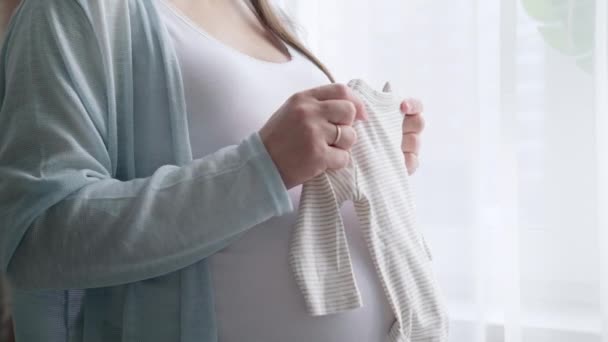 4k妊娠中の母親が赤ちゃんの服を手に持っている。幸せな期待の母親の子供を待っている。出産の準備,女の子の大きな腹の高度な妊娠. — ストック動画