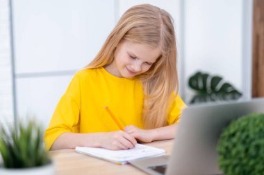 Evde ders çalışan sarışın kız dizüstü bilgisayarla okul ödevi yapıyor. Uzaktan öğrenme çevrimiçi eğitim. Okula geri dön.