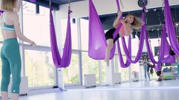 Молодая красивая женщина практикуется в аэрорастягивании качели. Упражнения аэродинамической йоги в фиолетовом гамаке в фитнес-клубе. Инструктор объясняет, как завершить упражнение — стоковое видео
