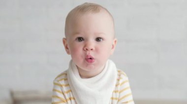 Evde portre yiyen bebek. Sağlıklı yemek, öğle yemeği zamanı. Çocuk komik suratlar yapıyor. Çocuk elinde kaseyle masada oturuyor.