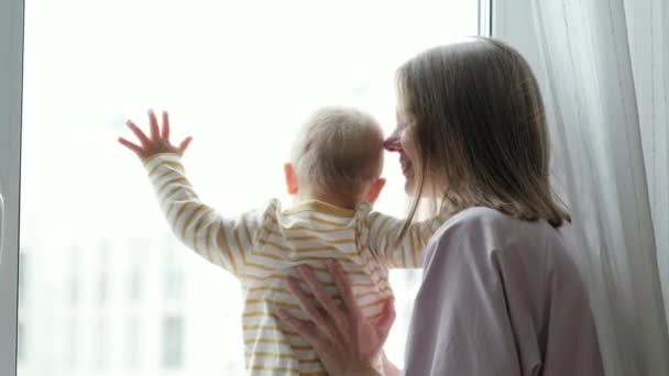 Jonge mooie moeder die haar kleine baby omarmt. Mam en dochter knuffelen elkaar zittend op de vensterbank en kijken door het raam. familie liefde. — Stockvideo
