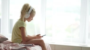 Kulaklıklı, elinde telefon olan küçük sarışın kız. Çocuk müzik dinleyerek eğleniyor. Dinlen, rahatla, evde kendine zaman ayır.