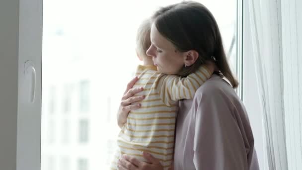 Küçük bebeğini kucaklayan genç ve güzel bir anne. Anne ve kızı birbirine sarılıyor pencere kenarında oturuyor ve pencereden dışarı bakıyor. Aile sevgisi. — Stok video