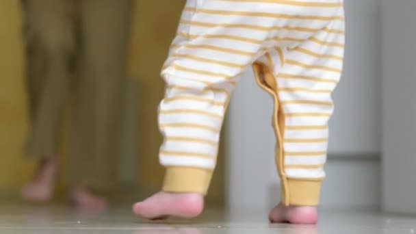Il bambino sta facendo i primi passi. Irriconoscibile bambina che cammina sul pavimento bianco a casa. Gambe scalze madre e bambino in cucina. — Video Stock