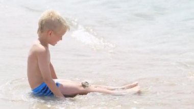 Kumsaldaki okyanusta kumla oynayan küçük sarışın çocuk. Aile tatilleri ve sıcak ülkelere seyahatler. Mutlu turizm tatili Mavi dalgalar suyu.