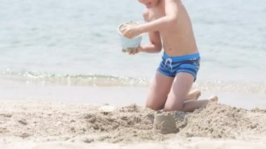 Kumsaldaki okyanusta kumla oynayan küçük sarışın çocuk. Çocuk kumdan kale inşa ediyor. Aile yaz tatilleri ve sıcak ülkelere seyahatler.