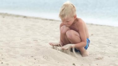 Kumsaldaki okyanusta kumla oynayan küçük sarışın çocuk. Aile tatilleri ve sıcak ülkelere seyahatler. Mutlu turizm tatili Mavi dalgalar suyu.