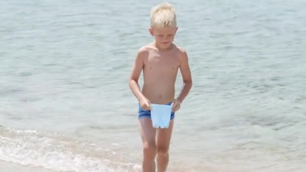 Kumsaldaki okyanusta kumla oynayan küçük sarışın çocuk. Çocuk kumdan kale inşa ediyor. Aile yaz tatilleri ve sıcak ülkelere seyahatler. — Stok video