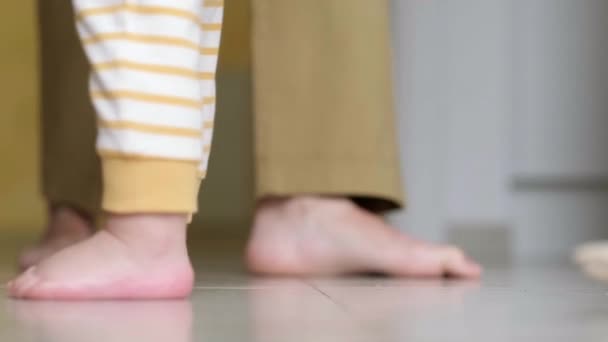 Petit garçon qui fait ses premiers pas. Petite fille méconnaissable marchant sur le sol blanc à la maison. pieds nus pieds mère et enfant sur cuisine. — Video
