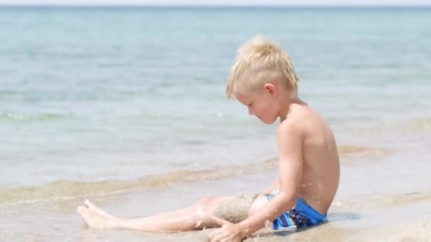 Malý blonďatý chlapec si hraje s pískem na pláži oceánu. Rodinné letní prázdniny a výlety do teplých zemí. Happy turistika dovolená modrá vlny voda. — Stock video