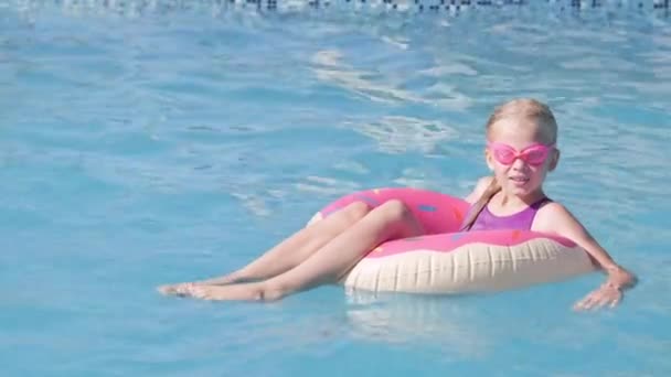 Retrato chica sonriente en la piscina, niño en gafas de baño sentado en anillo de natación rosado donut. Verano viajes familia hotel vacaciones turistas. — Vídeo de stock