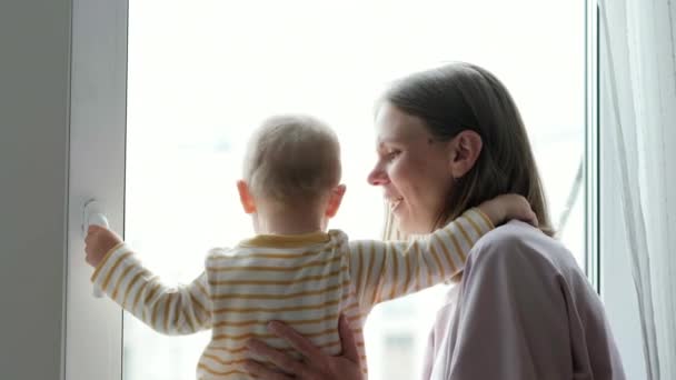 Joven hermosa madre abrazando a su pequeño bebé. Mamá e hija abrazándose juntas sentadas en el alféizar de la ventana y mirando por la ventana. amor familiar. — Vídeos de Stock