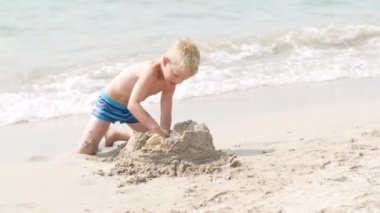 Kumsaldaki okyanusta kumla oynayan küçük sarışın çocuk. Çocuk kumdan kale inşa ediyor. Aile yaz tatilleri ve sıcak ülkelere seyahatler.