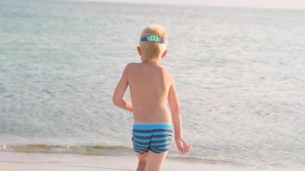 Вид ззаду щасливий маленький хлопчик біжить в океанську морську воду, щоб плавати. Літні канікули, відпустка, сімейна поїздка в теплі країни — стокове відео