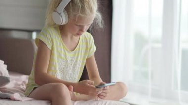 Kulaklıklı, elinde telefon olan küçük sarışın kız. Çocuk müzik dinleyerek eğleniyor. Dinlen, rahatla, evde kendine zaman ayır.