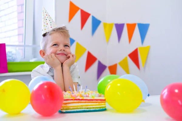 Blond kaukasisk pojke sitter omtänksamt och drömmande vid festligt bord nära födelsedag regnbåge kaka och gör en önskan. Färgglada bakgrund med ballonger — Stockfoto