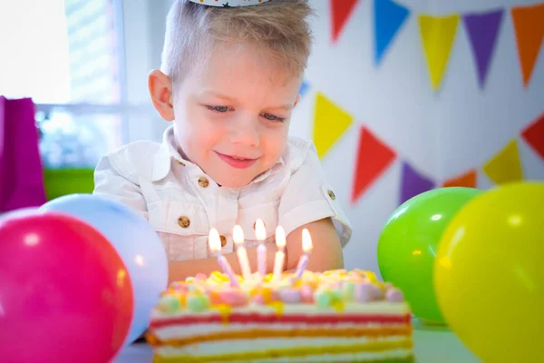 Rubia caucásica niño mirando velas en pastel de arco iris de cumpleaños, pidiendo un deseo antes de soplar hacia fuera en la fiesta de cumpleaños. Fondo colorido con globos —  Fotos de Stock