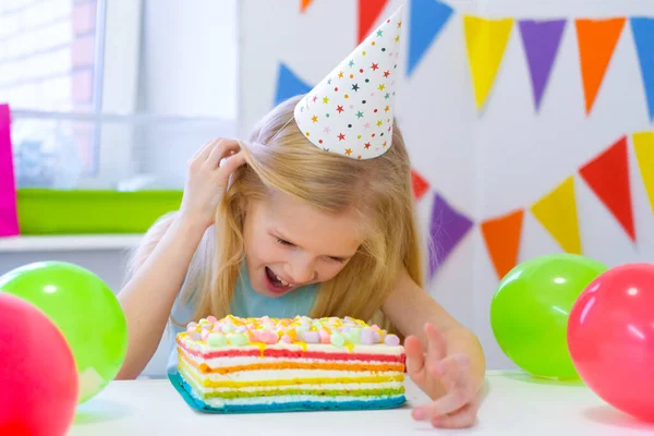 Blonde caucasian girl peeking out from behind birthday cake with a funny face on birthday party. Festive colorful background with balloons. Stock Picture