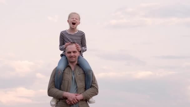 Joven papá alegremente pasar tiempo con su hija, jugando juntos al aire libre en verano. La muchacha encantadora se sienta sobre los hombros al padre y se ríe alegremente sobre el cielo nublado. día del padre. — Vídeos de Stock