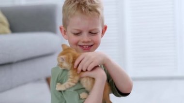 Küçük çocuk evde yerde kızıl kedi yavrusu kırmızı kurdelesiyle oynuyor. Çocukların ve hayvanların dostluğu, dostları ve kedilerin bakımı