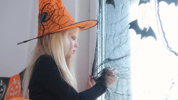 4k. Glad flicka i häxa kostym förbereder sig för Halloween Dekorera fönster i rummet av svarta fladdermöss — Stockvideo