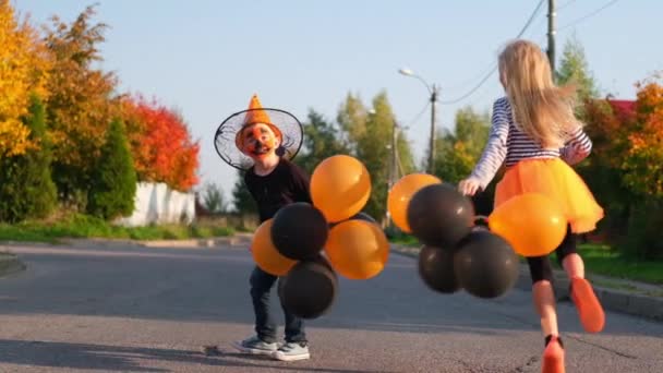 Langzame beweging. Halloween kinderen. Broer en zus in carnaval kostuums buiten. Jongen en meisje hebben plezier met oranje en zwarte ballonnen. — Stockvideo