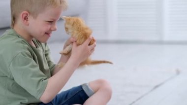 Küçük çocuk evde yerde kızıl kedi yavrusu kırmızı kurdelesiyle oynuyor. Çocukların ve hayvanların dostluğu, dostları ve kedilerin bakımı